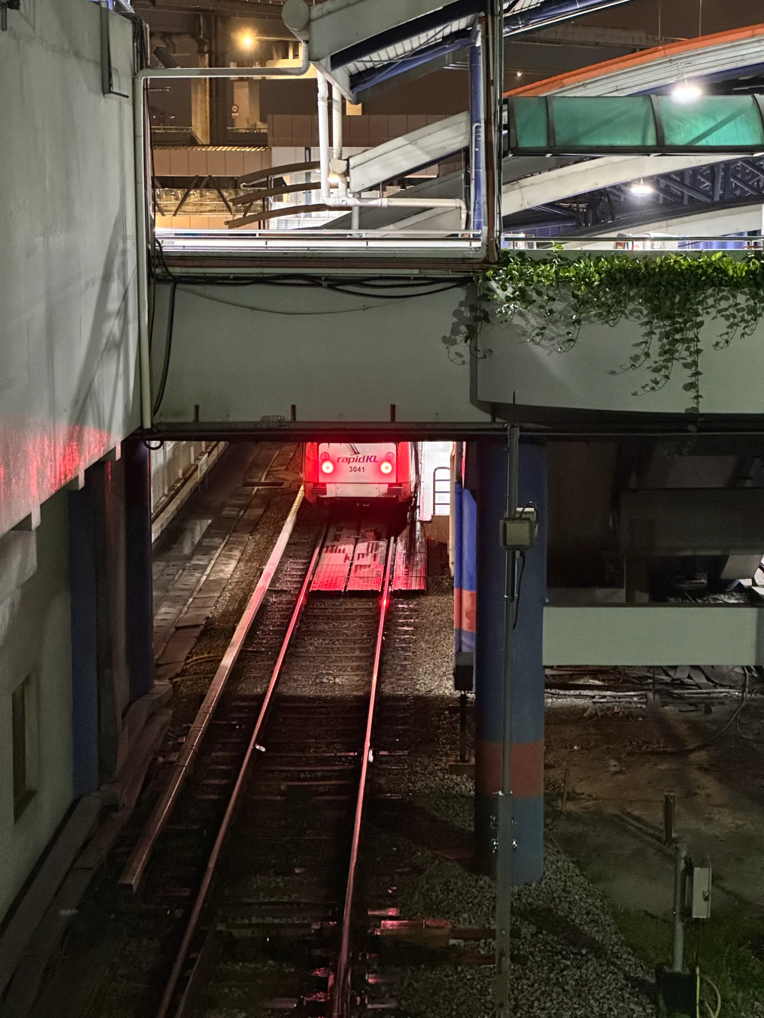 Looking down from a station footbridge at a rapidKL train parked under the slab, its red tail lights lighting the rails