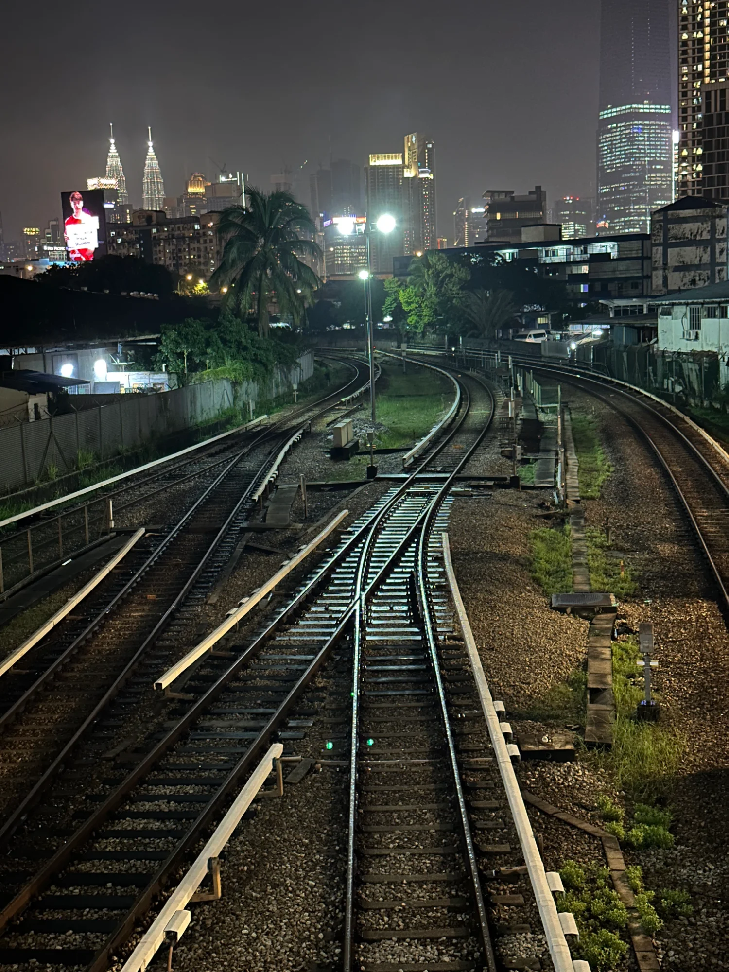 Rail tracks at night stretching from the bottom of the frame into the distance, with the Petronas Towers, Merdeka 118 and city lights